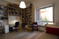 foto di una camera da letto singola, con libreria, scrivania, letto, poltroncina e finestra spalancata su un balcone