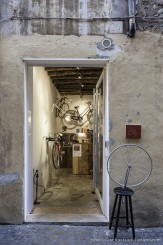 Veduta dall’esterno di Opera Unica, la porta di ingresso con accanto una copia della celebre opera di Duchamp “Ruota di Bicicletta”. La porta dello spazio è aperta e si intravede l’interno. 