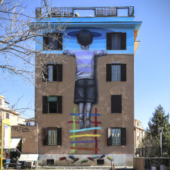 mural colorato sull’intera facciata della palazzina che rappresenta un bambino arrampicato su una scaletta colorata che con la testa sembra sfondare una superficie blu cielo